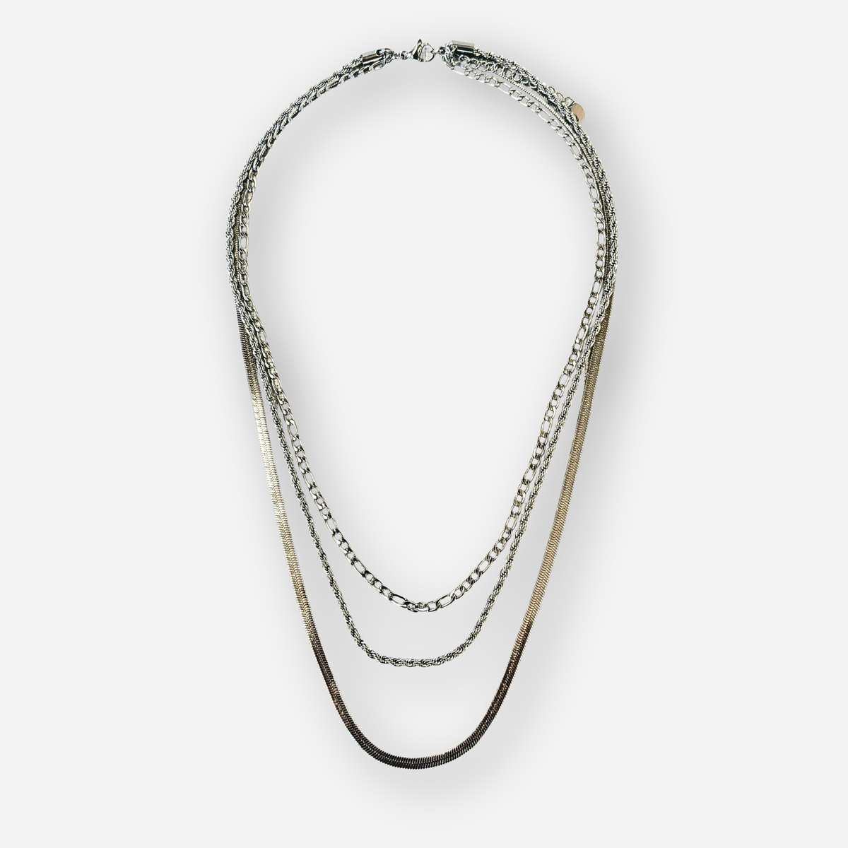 A layered necklace featuring three distinct chain styles in silver against a light gray background.