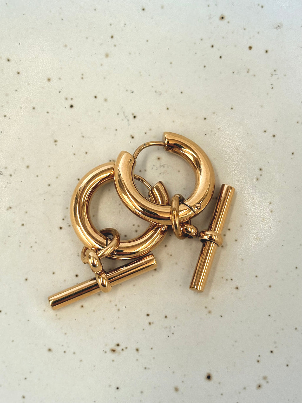 Gold hoop earring with a T bar on a light speckled background