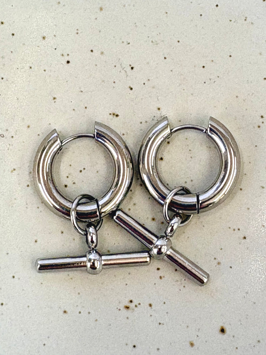 Pair of silver hoop earrings with T bar charms on a speckled surface