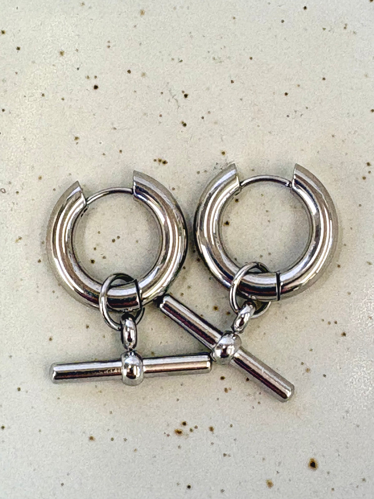 Pair of silver hoop earrings with T bar charms on a speckled surface