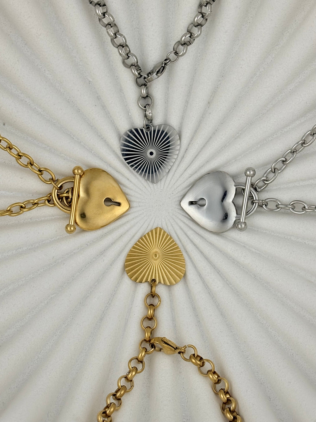 Four heart-shaped pendants on chains arranged in a circular pattern on a textured white background.