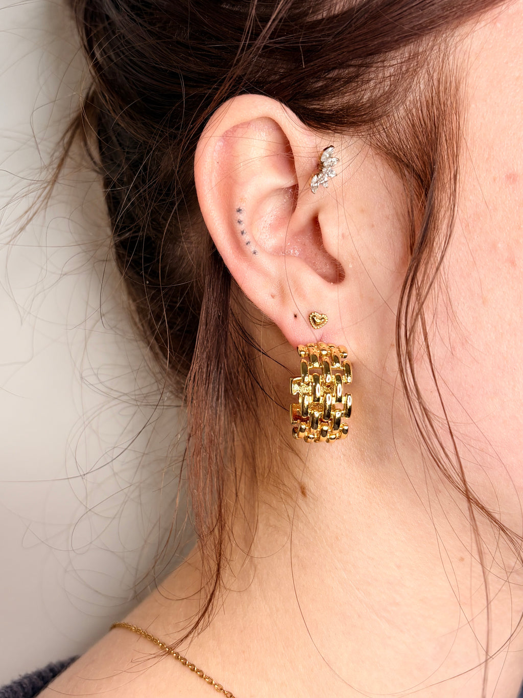 A close-up of a person's ear wearing a chunky gold hoop earring with a textured, woven-like chain design, along with other small ear piercings.