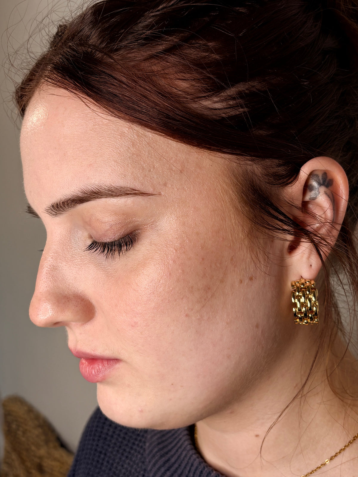 A close-up side profile of a woman with dark hair wearing a gold hoop earring with a textured, honeycomb-like pattern.