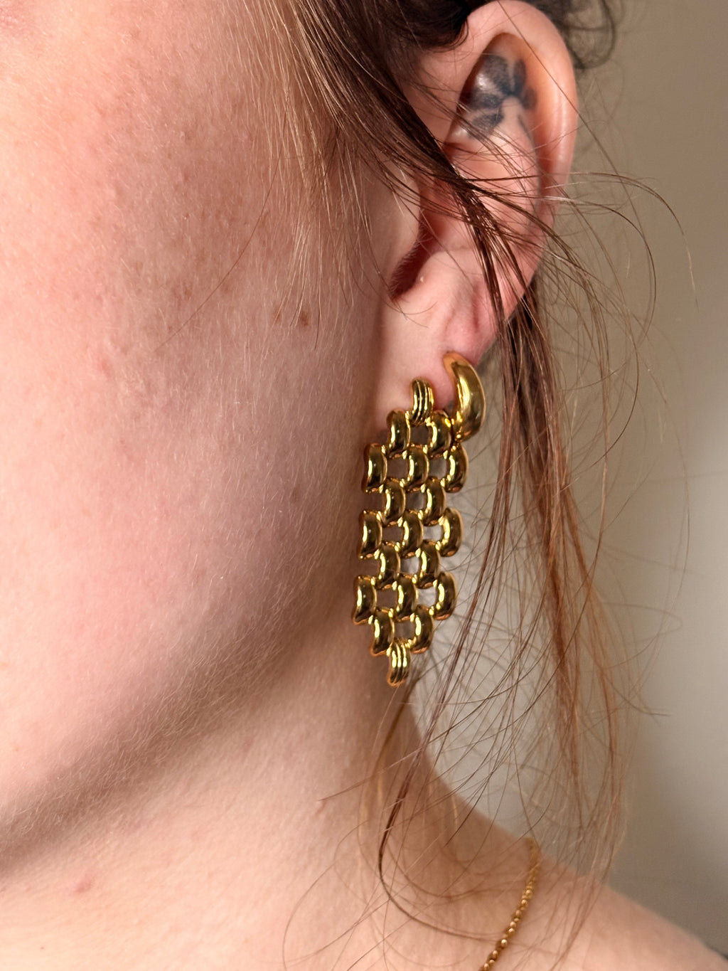 A close-up of a person's ear wearing a chunky, textured gold earring with a woven or chain-link style dangle.