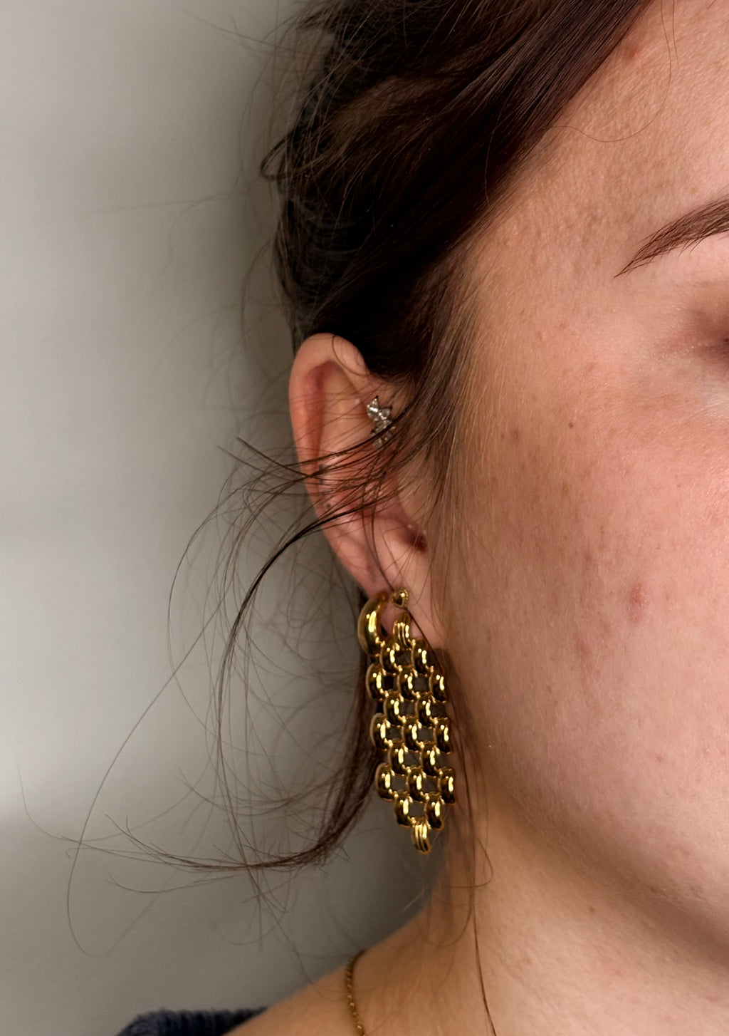 A close-up side profile of a woman wearing a chunky, gold-toned geometric dangle earring with a honeycomb-like textured pattern.