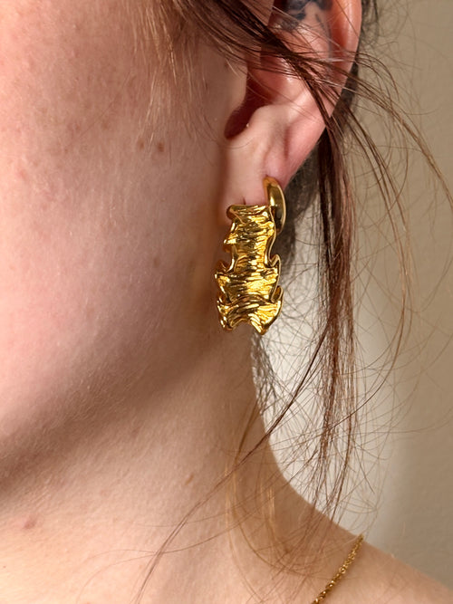 A close-up of a person's ear wearing a chunky, textured gold hoop earring.