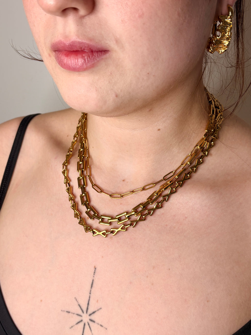 A close-up of a model wearing a layered gold chain necklace with chunky links, paired with a black spaghetti strap top.