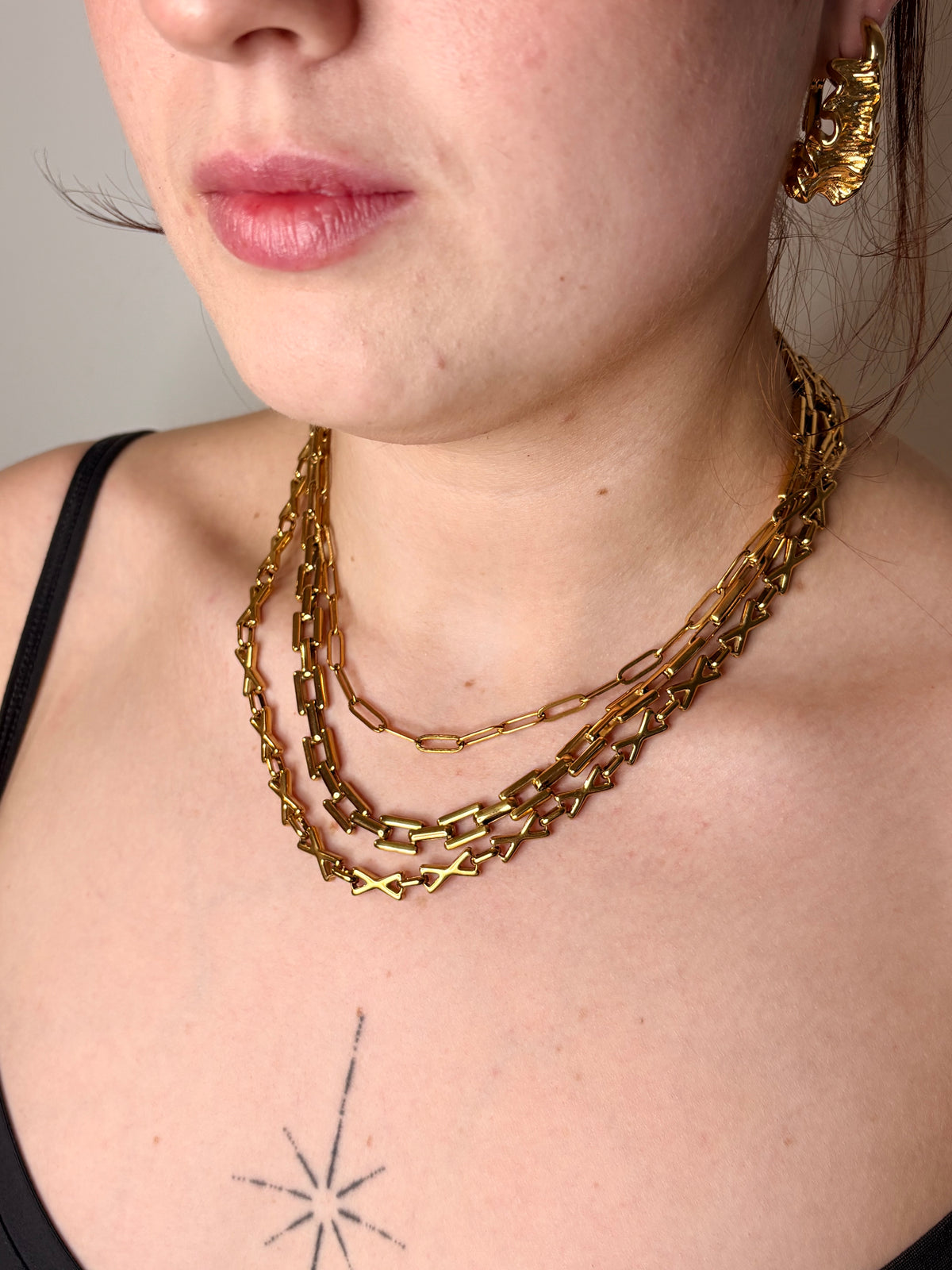 A close-up of a model wearing a layered gold chain necklace with chunky links, paired with a black spaghetti strap top.