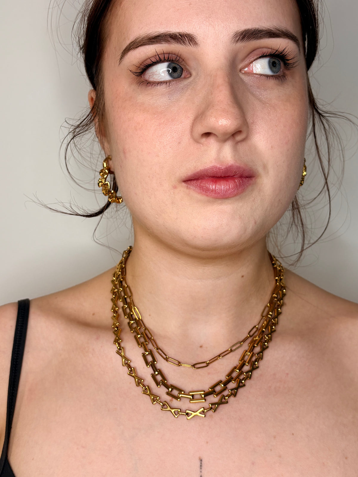 A close-up of a woman wearing a multi-layered gold chain necklace and matching gold hoop earrings against a plain light gray background.