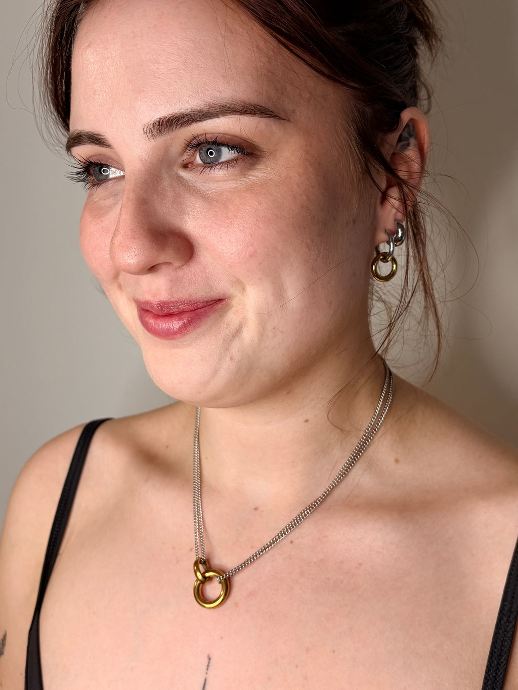 A close-up portrait of a woman wearing a gold ring pendant necklace on a silver chain, along with matching gold hoop earrings, against a plain neutral background.
