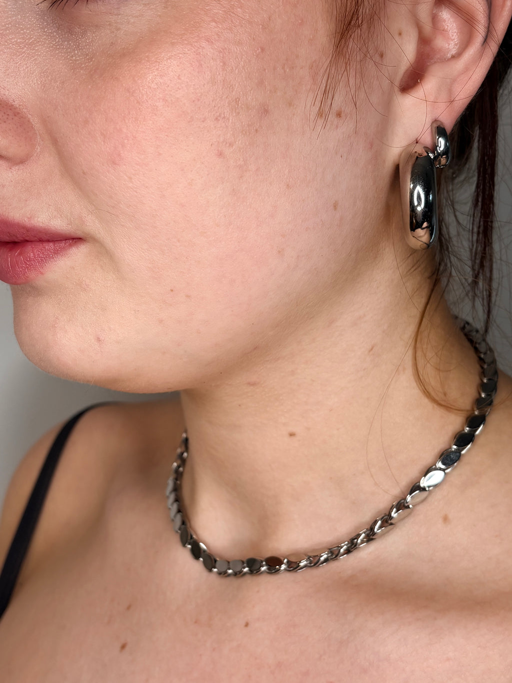 A close-up of a person's neck and lower face wearing a chunky silver chain necklace and a matching silver hoop earring.