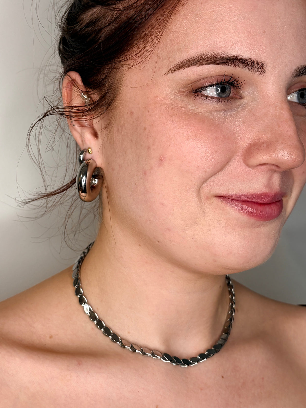 A close-up of a woman wearing a chunky silver chain necklace and matching silver hoop earrings.