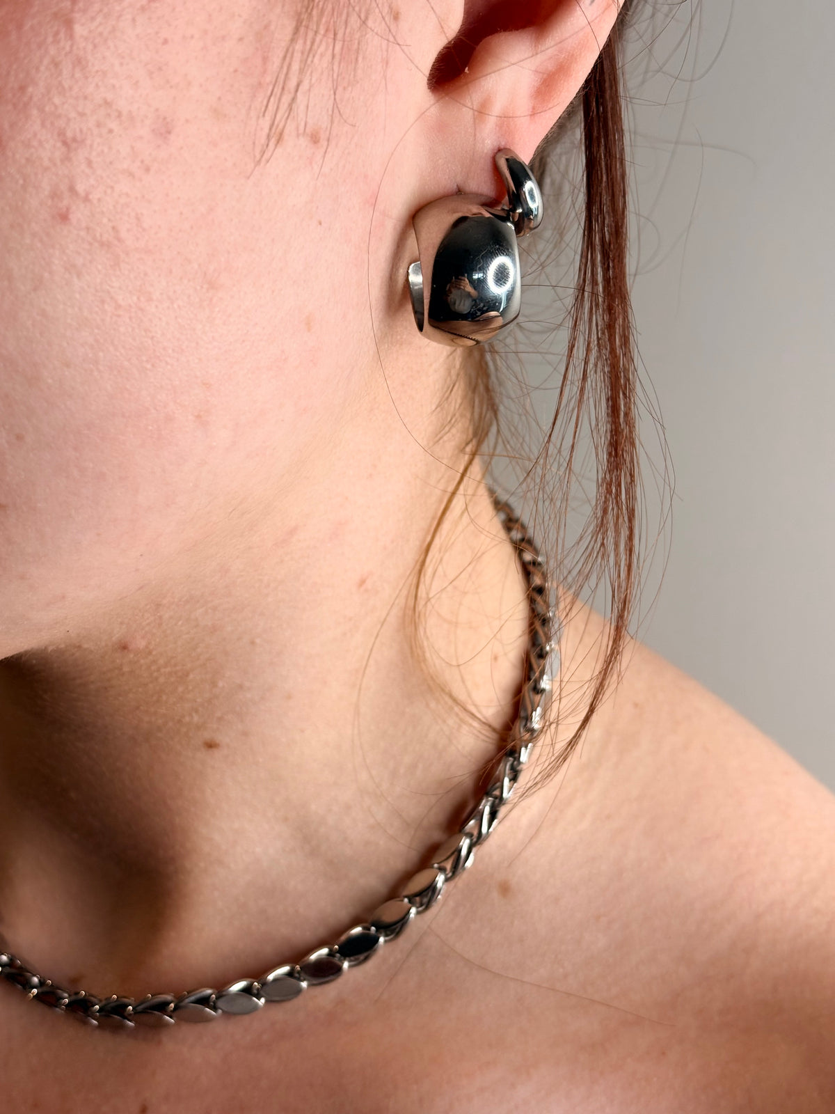 A close-up of a person's ear wearing a polished silver crescent-shaped earring, with a matching silver chain necklace also visible against a plain light gray background.