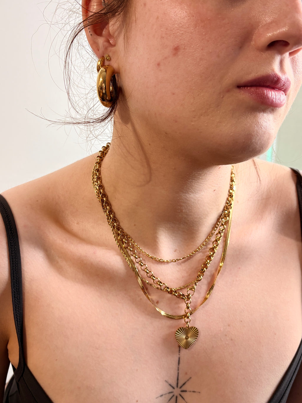 A close-up of a person wearing layered gold necklaces, including a textured chain, a thin chain with a small heart charm, and a gold hoop earring, against a plain light background.