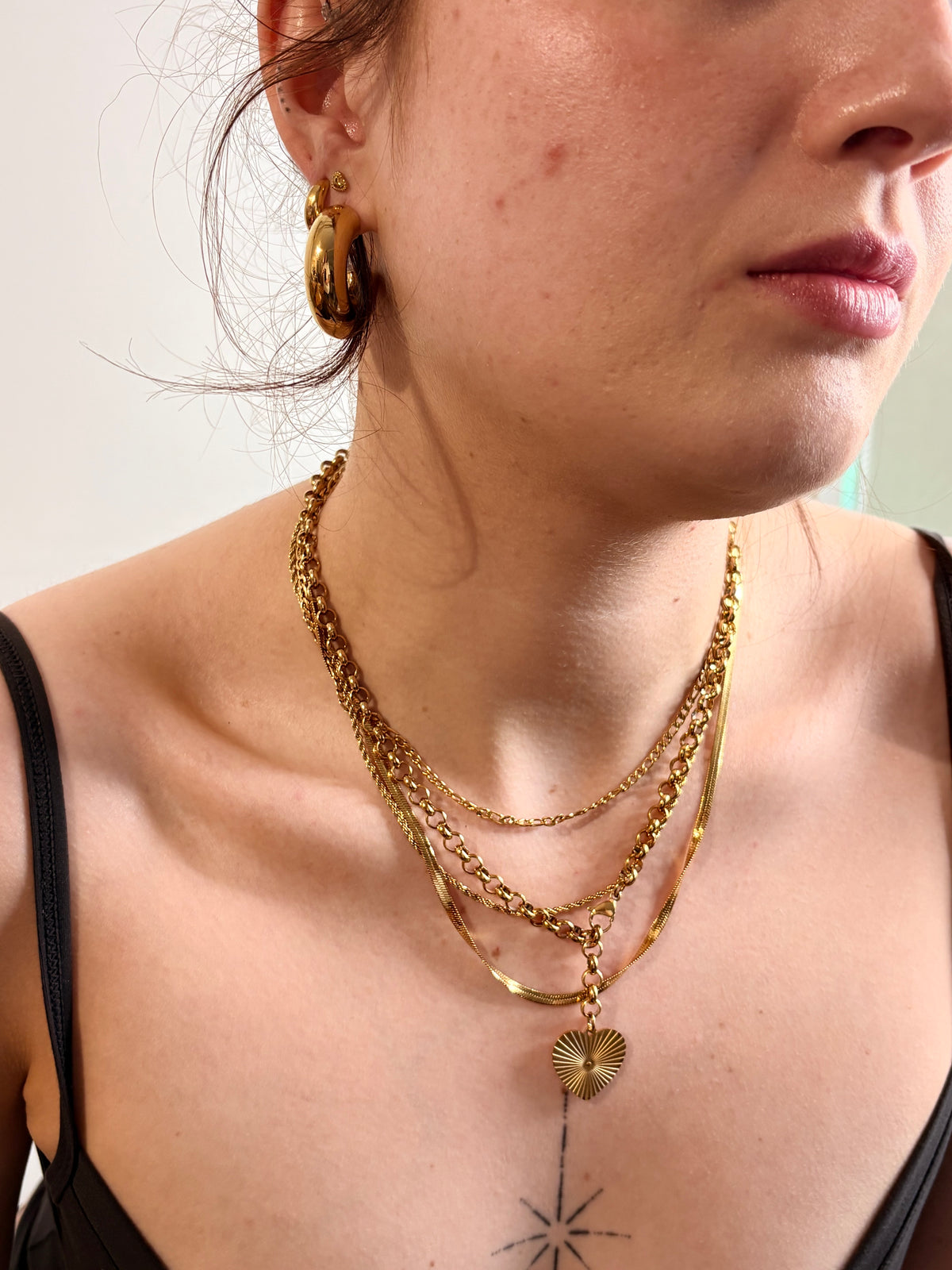 A close-up of a person wearing layered gold necklaces, including a textured chain, a thin chain with a small heart charm, and a gold hoop earring, against a plain light background.